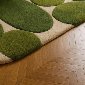 May include: A close-up of a rug with a beige background and green, organic-shaped patterns. The rug is placed on a wooden floor with a herringbone pattern. The rug's design resembles stepping stones.