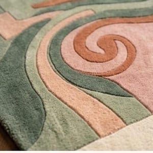 May include: Close-up of a colorful rug with an abstract design. The rug features a mix of colors, including green, peach, and brown, arranged in flowing, curved patterns. The texture appears soft and plush, suggesting a comfortable floor covering.