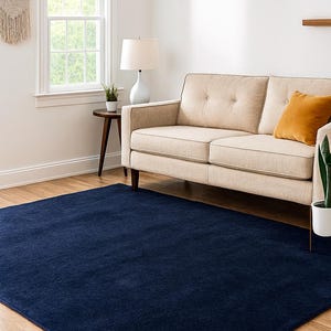 May include: A navy blue rectangular rug on a hardwood floor. The rug is in a living room setting, with a beige sofa, a side table with a lamp, and a window in the background. The rug is a solid color and covers a large area of the floor.