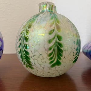May include: A white glass vase with green and iridescent designs. The vase is round and has a small opening at the top.