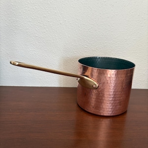 May include: A copper saucepan with a hammered finish and a brass handle. The interior of the pan is a dark teal color. The handle extends horizontally from the side of the pan.
