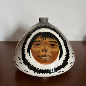 May include: A white ceramic vase with a rounded, triangular shape. The vase is decorated with a black and white illustration of a person wearing a hooded garment. The person's face is painted in brown, black, and white.