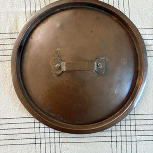May include: A round, copper-colored pot lid with a handle. The lid has a slightly raised rim and a small, rectangular handle attached with rivets. The surface shows signs of age and use, with variations in color and texture.