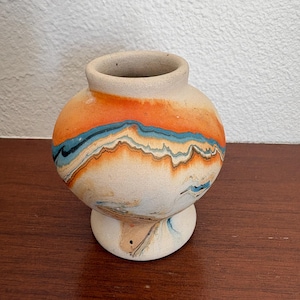 May include: A small, round ceramic vase with a wide, open mouth and a pedestal base. The exterior features a marbled pattern with horizontal bands of bright orange, light blue, and cream, creating a fluid, layered effect across the surface.
