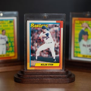 May include: A baseball card featuring Nolan Ryan in a Rangers uniform, displayed in a protective case and wooden stand. The card has a colorful design with the team logo and player's name. Two other cards are visible in the background.