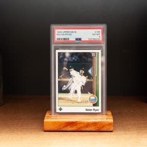 May include: A baseball card of Nolan Ryan from the 1989 Upper Deck series, graded EX-MT, displayed in a clear protective case. The card is set on a small, rectangular wooden stand. The background is a dark surface.