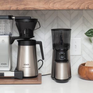May include: A stainless steel coffee maker and grinder set with a wooden base and a wooden bowl. The coffee maker has a black top and a silver carafe. The grinder is black and silver.