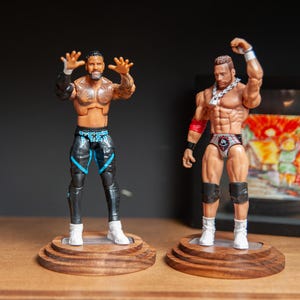 May include: Two action figures of muscular men on wooden bases. One wears black and blue wrestling gear, the other wears red and silver trunks. Both figures have their arms raised in a pose.