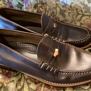 May include: A pair of dark brown leather penny loafers with white stitching and a decorative strap. The shoes have a classic design and are set against a floral background.