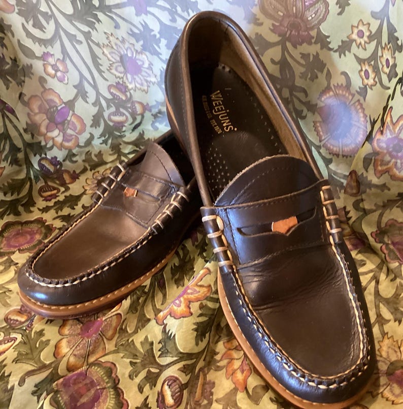 May include: A pair of dark brown leather Weejuns penny loafers with white stitching and a light brown sole. The shoes are displayed on a floral patterned fabric.