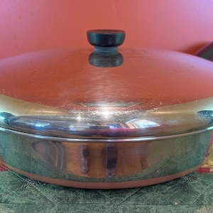 Puede incluir: Una olla de cocina de acero inoxidable de color cobre con tapa. La tapa tiene un asa negra y un pomo negro. La olla está sobre una superficie estampada.