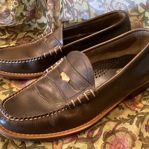 May include: A pair of dark brown leather penny loafers with white stitching around the soles and uppers. The shoes have a classic design with a gold-colored accent on the vamp. The soles are a lighter brown color.