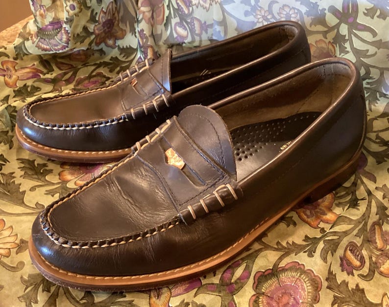May include: A pair of dark brown leather penny loafers with white stitching around the soles and uppers. The shoes have a classic design with a gold-colored accent on the vamp. The soles are a lighter brown color.