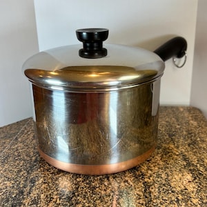 May include: A stainless steel saucepan with a copper base and a matching lid. The pot features a long black handle with a metal hanging loop at the end and a black knob on the center of the lid. The exterior surface shows light surface scratches.