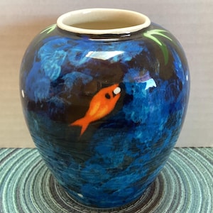 Puede incluir: Un jarrón de cerámica de forma redonda, que representa una escena submarina. El jarrón es principalmente azul con un pez naranja pintado. El borde es blanquecino, y el diseño incluye detalles en azul oscuro y negro.