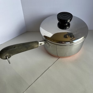 Pode incluir: Uma panela de aço inoxidável com tampa e cabo de madeira. A panela tem uma base de cor cobre e um botão preto na tampa. A alça tem um laço de metal na extremidade.