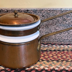 May include: A copper double boiler with a lid and long handle. The pot has a copper finish with white and copper inserts. The handle is a brass color. The pot is sitting on a patterned surface.