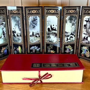 May include: A white ceramic mug with a black handle and the text "But First, Coffee" in a bold, black font. The mug is set against a blurred background, suggesting a cozy setting. The mug appears to be approximately 4 inches tall.