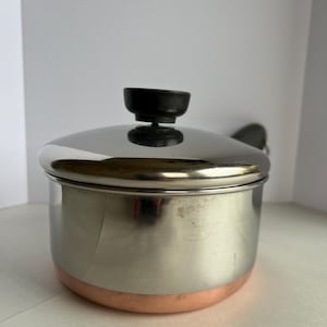 Puede incluir: Una cacerola de acero inoxidable con base de color cobre y tapa a juego. La tapa tiene un asa negra. La olla está diseñada para cocinar y es adecuada para diversas tareas de cocina.