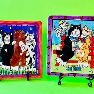 以下が含まれることがあります： 猫のイラストが描かれた2つの正方形のセラミックプレート。1つのプレートには、星空の下で抱き合う猫が描かれています。もう1つのプレートには、花を持つ猫がベンチに座っている様子が描かれています。どちらのプレートもカラフルなボーダーがあり、黒いスタンドに飾られています。