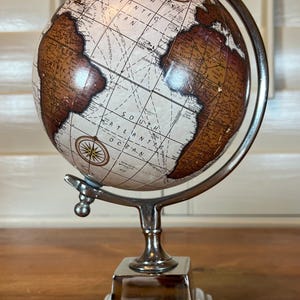May include: A decorative globe with a cream-colored surface and brown continents. The globe is mounted on a silver-colored metal stand with a square base. Compass rose details are visible on the globe's surface.
