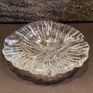 Puede incluir: Un cuenco de vidrio transparente con un diseño texturizado y orgánico. El cuenco tiene un borde festoneado y un interior ondulado, creando una apariencia artística única. El cuenco es probablemente para fines decorativos.