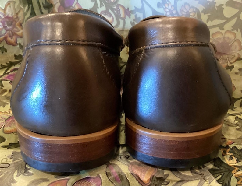 May include: A pair of brown leather loafers, viewed from the back. The shoes have a polished, glossy finish and a light brown sole. The stitching is visible along the top of the shoe.
