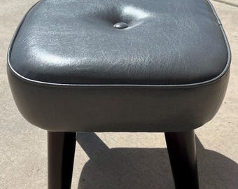 Small square foot stool in grey vinyl or faux leather.  Unknown brand but well-made & like new. MCM with 4 dark wood legs. 12.5x12.5x11 in.