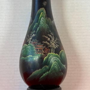Puede incluir: Jarrón negro con una escena de paisaje pintada que presenta montañas verdes, árboles y un edificio tradicional. El jarrón está montado sobre un soporte negro decorativo. El jarrón tiene un cuello estrecho y un cuerpo bulboso.