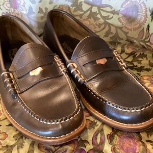 May include: A pair of dark brown leather penny loafers with white stitching and a light brown sole. The shoes have a classic design with a strap across the vamp and a small decorative accent.