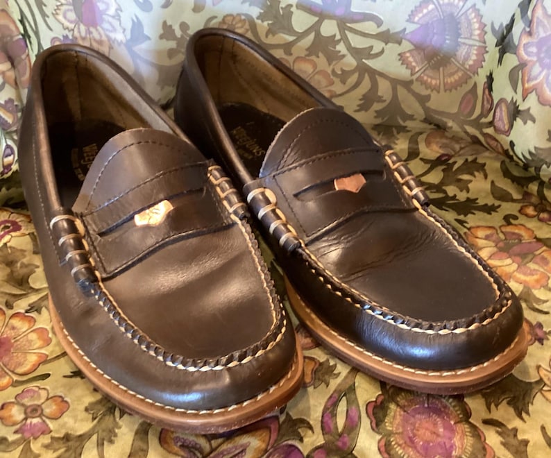May include: A pair of dark brown leather penny loafers with white stitching and a light brown sole. The shoes have a classic design with a strap across the vamp and a small decorative accent.