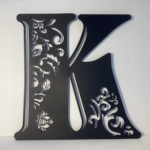Letra "K" colgante recortada con encaje metálico. De color negro oscuro y elegante. Usada, con ligeras señales de uso. 33 x 33 x 12,7 cm.