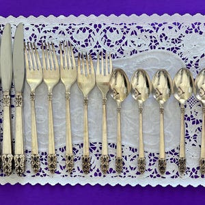 Puede incluir: Una colección de cubiertos antiguos, que incluye cuchillos, tenedores y cucharas, dispuestos sobre un mantel individual de encaje blanco sobre un fondo púrpura. La cubertería tiene mangos ornamentados y un acabado pulido.