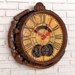 May include: A round wall clock with a dark brown, weathered metal frame and a textured, light tan face. It features black Roman numerals, bright red clock hands, and two visible mechanical gears at the bottom. The design resembles a vintage pocket watch.