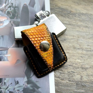 Puede incluir: Un estuche de cuero hecho a mano para encendedor con un patrón de piel de reptil en tonos naranja y amarillo. El estuche es negro en los laterales y tiene un cierre de botón plateado. Un encendedor plateado es parcialmente visible en el fondo.