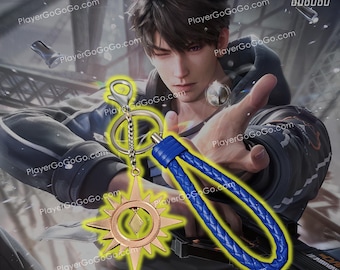 May include: A gold-coloured keychain with a star-shaped charm and a blue braided strap. The charm has a diamond-shaped design in the centre. The keychain is attached to a silver key ring.