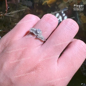 Op de afbeelding: Een zilveren ring met een grote, heldere edelsteen in het midden, omringd door kleinere stenen en een bladvormig ontwerp. De ring bevindt zich op een vinger en toont de ingewikkelde details en schittering.