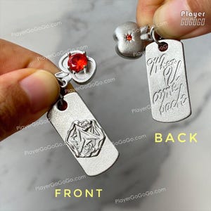 May include: Two silver-toned metal pendants featuring a red gemstone set in an apple-shaped topper. One pendant shows a hexagonal embossed design on the front, while the other displays the engraved text "When U come back" on the back.