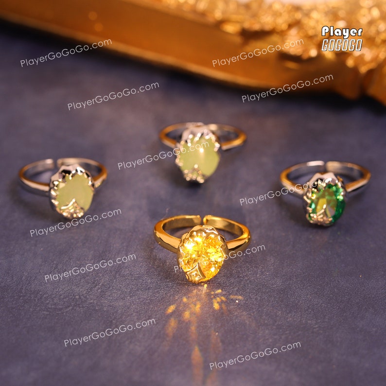May include: Four gold-tone rings with different colored stones. The stones are green, yellow, and light green. The rings are on a gray surface.