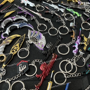 Op de afbeelding: Een verzameling kleurrijke metalen sleutelhangers met verschillende wapenontwerpen, waaronder zwaarden, messen en pistolen. De sleutelhangers zijn bevestigd aan zilveren sleutelringen en kettingen. De sleutelhangers worden gepresenteerd op een zwart oppervlak. PlayerGoGoGo.com is op verschillende sleutelhangers gedrukt.