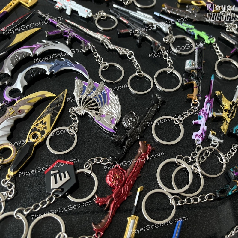 K&ouml;nnte beinhalten: Eine Sammlung bunter Schl&uuml;sselanh&auml;nger aus Metall mit verschiedenen Waffendesigns, darunter Schwerter, Messer und Pistolen. Die Schl&uuml;sselanh&auml;nger sind an silbernen Schl&uuml;sselringen und Ketten befestigt. Die Schl&uuml;sselanh&auml;nger werden auf einer schwarzen Oberfl&auml;che pr&auml;sentiert. PlayerGoGoGo.com ist auf mehreren der Schl&uuml;sselanh&auml;nger aufgedruckt.
