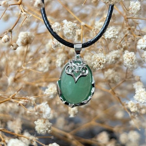 Pode incluir: Um colar com pingente de prata com uma pedra preciosa oval verde. O pingente tem uma moldura de prata decorativa e pende de um cordão preto. O fundo é um foco suave de flores secas.