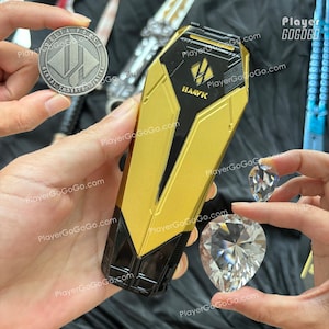 Könnte beinhalten: Ein gold-schwarzes rechteckiges Gehäuse mit dem Aufdruck "HAAVK". Zwei klare, facettierte, tropfenförmige Edelsteine werden in der Nähe gehalten. Eine Silbermünze mit der Aufschrift "DELTA FORCE" ist ebenfalls zu sehen.