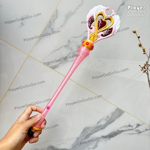 May include: A pink and gold toy wand with a heart-shaped design at the top, featuring white wings and a gold heart. The wand has a long, slender pink handle with a few pink gems. The text "Player GoGoGo" is visible.