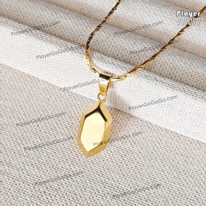 Könnte beinhalten: Eine goldfarbene Halskette mit einem geometrischen Anhänger. Der Anhänger hat ein facettiertes Design und hängt an einer zarten Kette. Der Hintergrund ist ein strukturierter, cremefarbener Stoff.