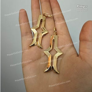 May include: A pair of gold-colored earrings with a unique, geometric design. Each earring features a stylized star shape with curved edges and a hook closure. The earrings are displayed against a neutral background.