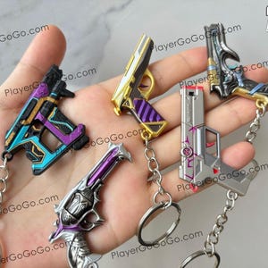 K&ouml;nnte beinhalten: Ein Set mit f&uuml;nf pistolenf&ouml;rmigen Schl&uuml;sselanh&auml;ngern. Die Schl&uuml;sselanh&auml;nger sind aus Metall und haben verschiedene Designs und Farben, darunter Silber, Gold, Lila und Blau. Die Schl&uuml;sselanh&auml;nger sind an silbernen Ketten befestigt.