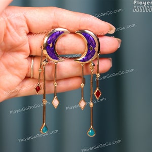 Puede incluir: Un par de pendientes dorados con forma de media luna con esmalte morado y colgantes. Los colgantes incluyen gemas rojas, rosas y turquesas. Los pendientes están sostenidos por una mano.