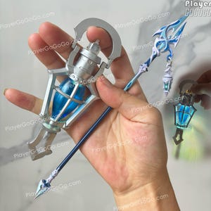 May include: A silver-toned metal staff accessory featuring a central lantern-shaped component with a bright blue translucent core. The staff is topped with a decorative blue and white trident-like finial and includes a small hanging lantern charm.