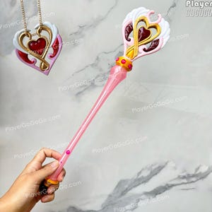Könnte beinhalten: Ein rosafarbener Kunststoffstab mit herzförmigem Kopf, weißen Flügeln und goldenen Akzenten, dazu ein passender herzförmiger Anhänger an einer goldenen Kette. Der Stabgriff ist transparent rosa mit kleinen rosa kreisförmigen Details und einem Goldring.